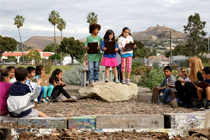 A group of children holding papers presenting to classmates at an outdoor learning classroom at will Rogers Two-Way Immersion School of Environmental Science.